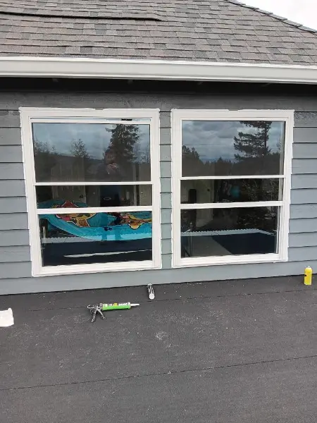Pair of new white double-hung windows on blue siding with a caulk gun on the roof below