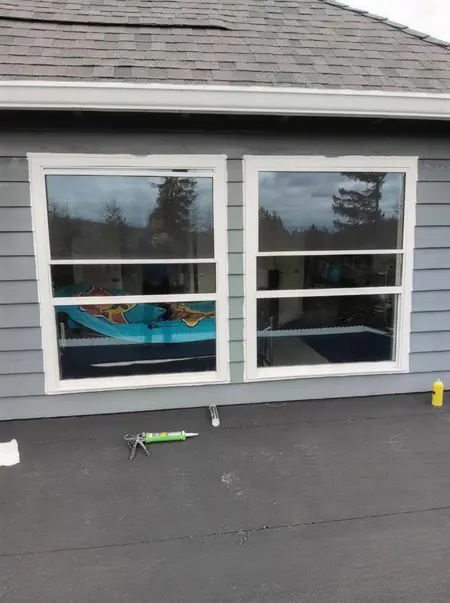 Pair of new white double-hung windows on blue siding with a caulk gun on the roof below