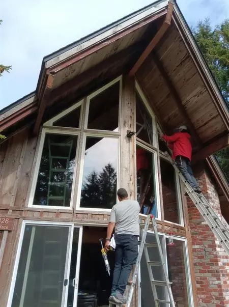 Three installers on ladders setting large multi-pane windows in the gable of a wood cabin-style house