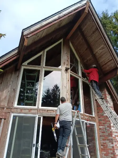 Three installers on ladders setting large multi-pane windows in the gable of a wood cabin-style house