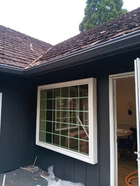 Large grid picture window on a charcoal house corner under a worn cedar shake roof, door open mid-job