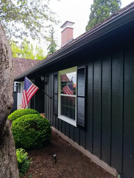 Window with black shutters on a dark green house beside American flags and a boxwood shrub