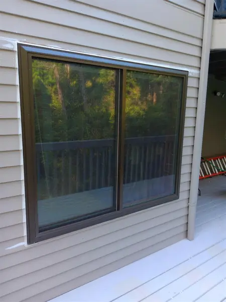 Dark bronze-framed sliding window on tan lap siding beside a downspout, above a painted deck