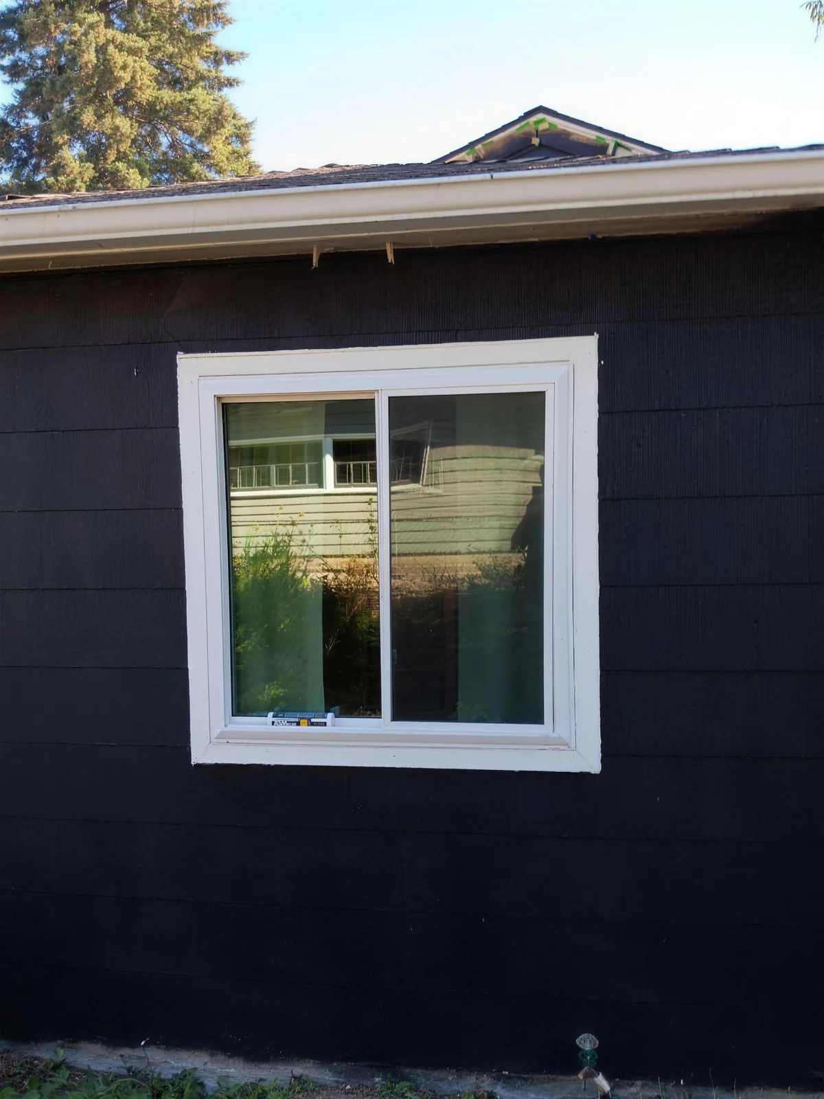 White-trimmed square sliding window centered on a dark-painted wall beneath a roof overhang, with a tall evergreen behind