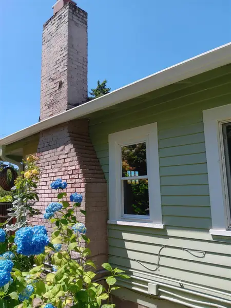 White-trimmed window beside a brick chimney, green siding and blue hydrangeas