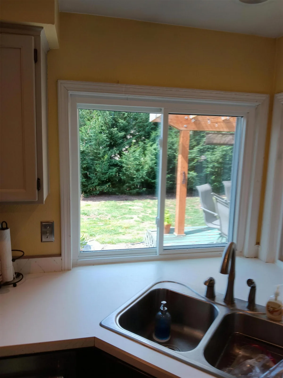 White-framed sliding window above a kitchen sink on a yellow wall, looking out to a green lawn and a wood pergola
