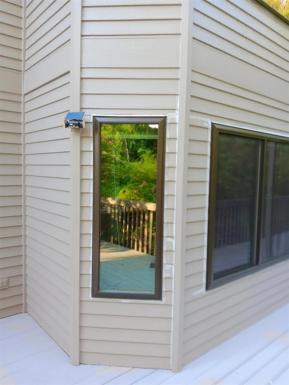 Tall narrow dark-framed window at the corner of a tan lap-siding house, beside a wider sliding window and a white deck