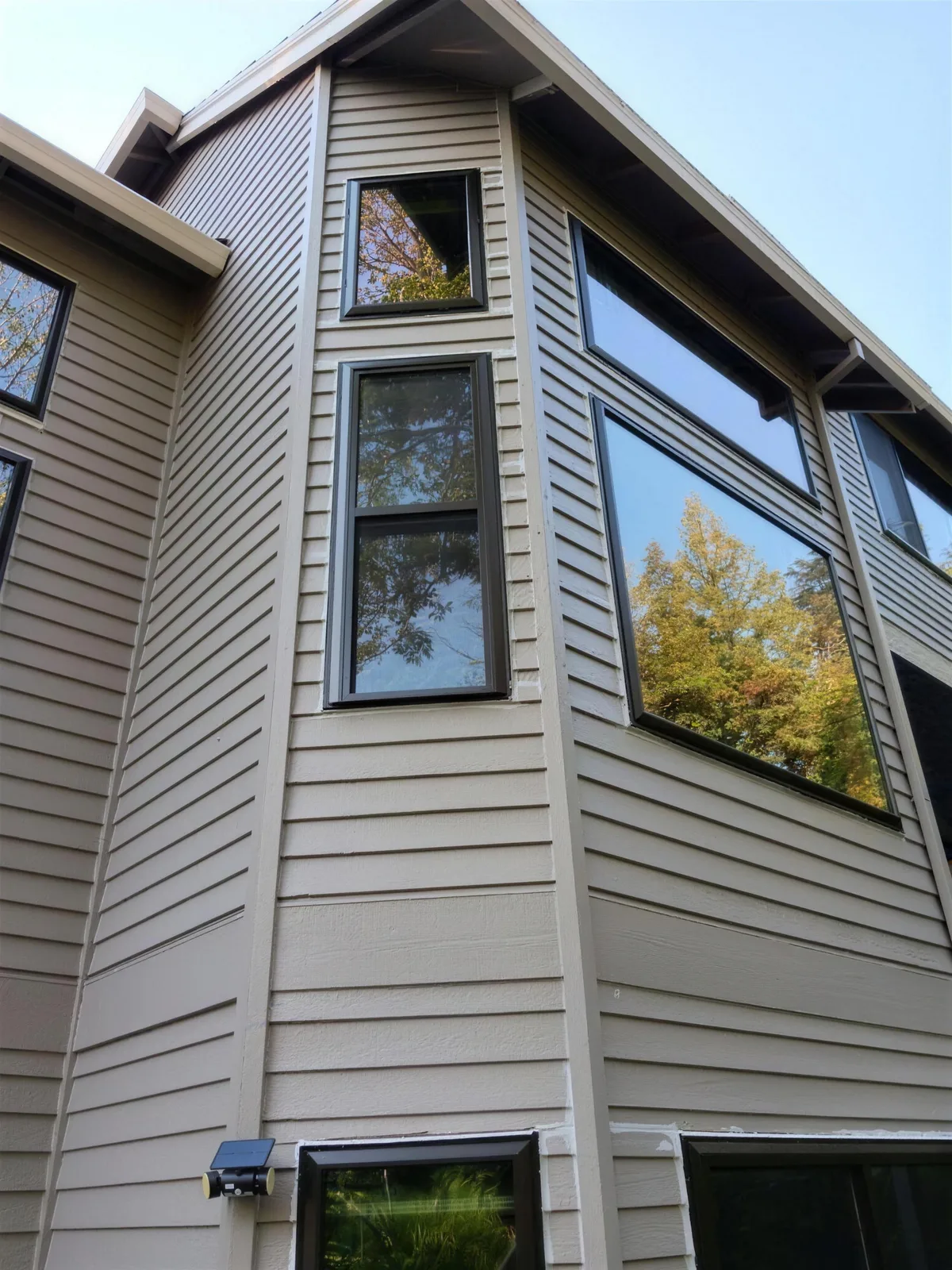 Looking up at black-framed windows on a tall tan lap-siding wall, a two-part window above a large picture window, under a blue sky