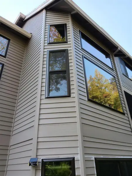 Looking up at black-framed windows on a tall tan lap-siding wall, a two-part window above a large picture window, under a blue sky