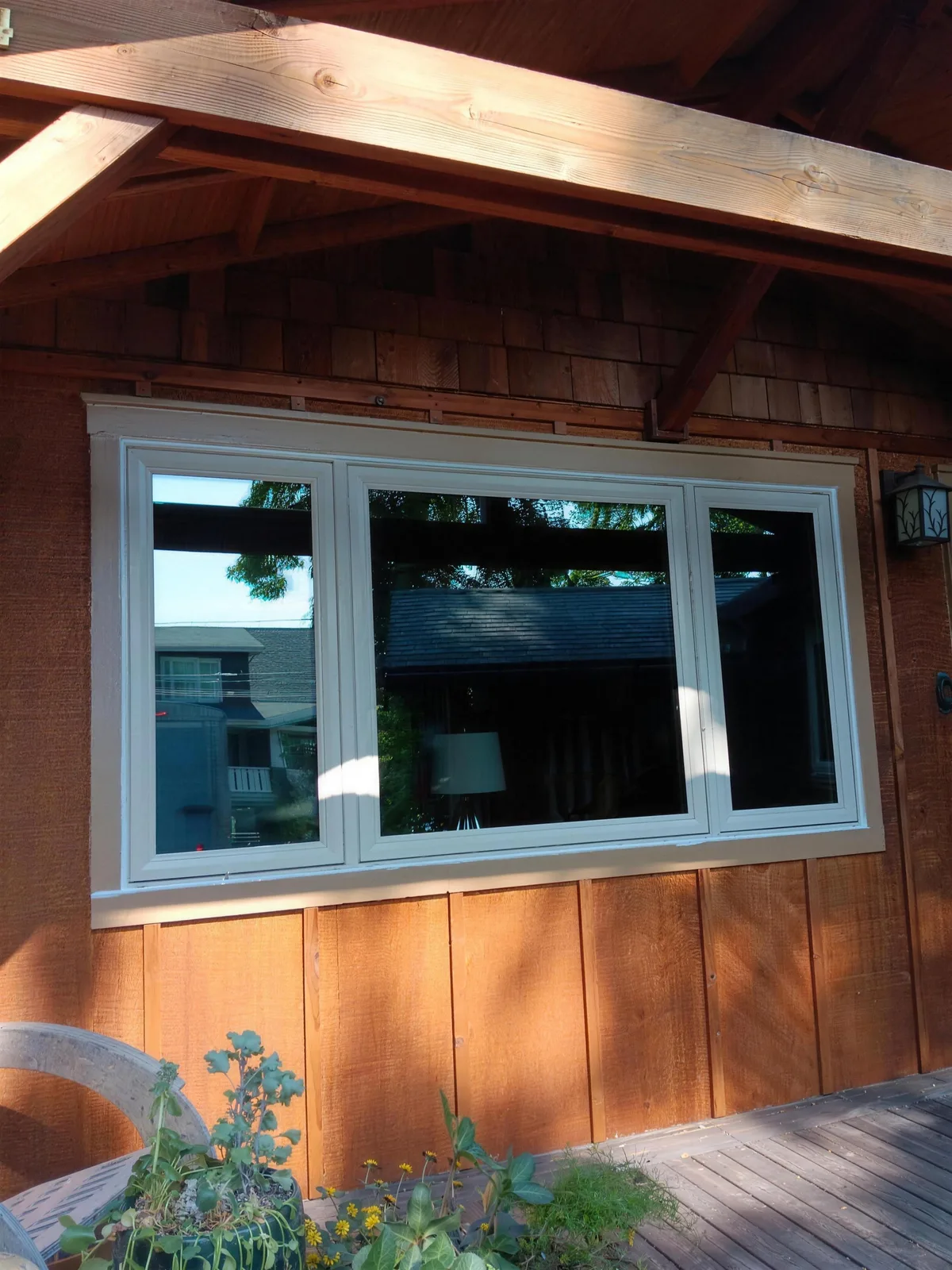 Wide white three-panel window beneath an exposed timber roof on a wood-sided home