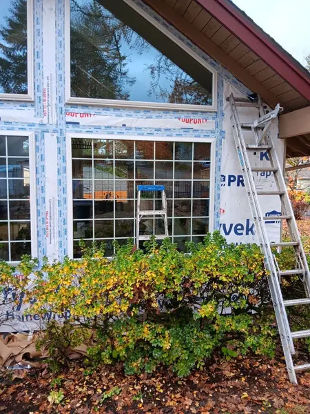 Wall wrapped in house wrap with wide flashing tape around a triangular gable window and a large gridded window, a ladder and a shrub in front