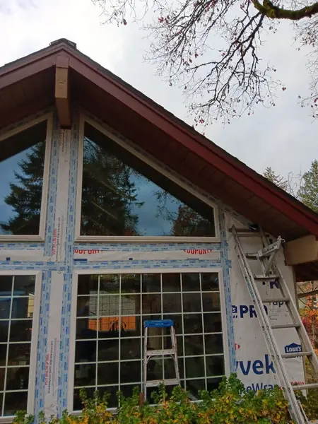 Gable end of a house with two triangular windows and gridded windows below, all taped with flashing over house wrap, a ladder against the wall
