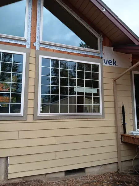 Triangular gable windows with blue flashing tape above a large white window with grid bars and tan trim, house wrap still exposed beside the lap siding