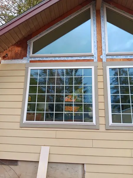 Two triangular gable windows with flashing tape above two large gridded windows with tan trim, on a house with tan lap siding going up