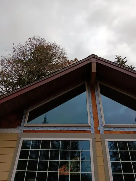 Looking up at two large triangular gable-end windows with white frames, taped with blue flashing around the openings, under a dark red roof edge