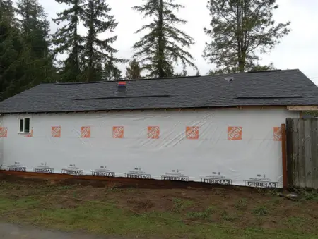 Long garage-style building wrapped in white house wrap under a new dark shingle roof, with one small window