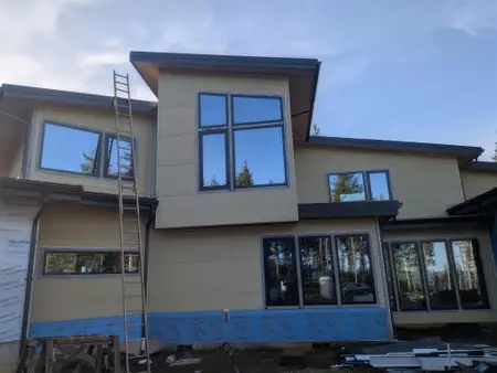 Two-story house with large tan wall panels and horizontal lap siding around black-framed windows, blue flashing membrane along the base and a ladder against the wall