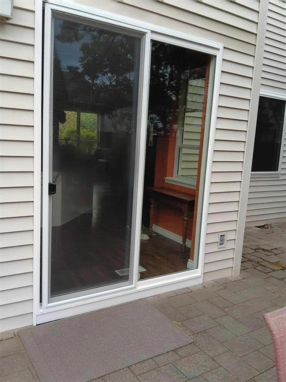 Sliding glass door with a white frame and one panel slid partly open, on a tan lap-siding house above a paver patio