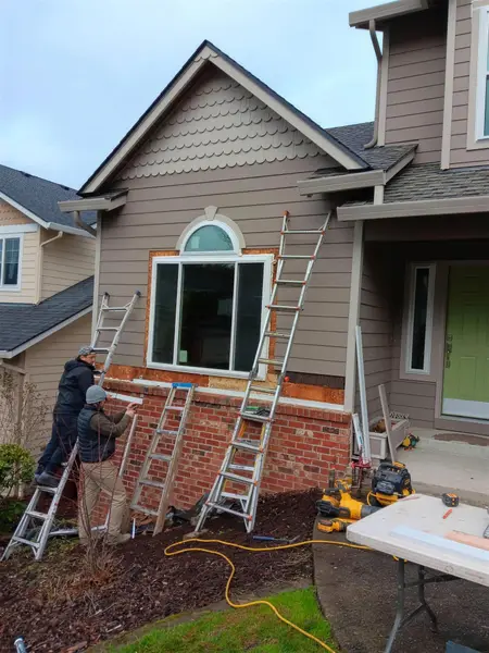 Two installers on ladders trimming out a new picture window with an arched transom on a taupe two-story house with a brick base, bare sheathing still exposed around the opening